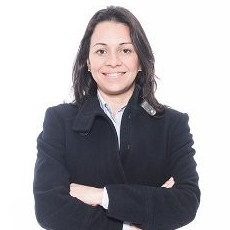 Mulher sorrindo com jaqueta preta, braços cruzados.