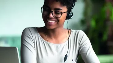 Mulher sorrindo, usando fones de ouvido e olhando para laptop.