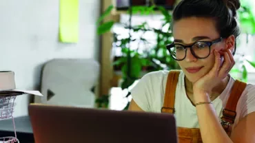 Mulher com óculos trabalha no laptop em uma mesa com plantas ao fundo.