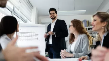 Executivo em pé apresentando gráficos para grupo em reunião.