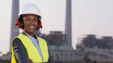 Mulher preta usando equipamentos de proteção na frente de uma indústria