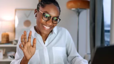 Mulher de pele negra e cabelo preto trançado acena para o computador enquanto está em uma reunião de trabalho.