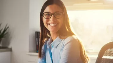 Pessoa sorrindo sentada em escritório com luz solar ao fundo.