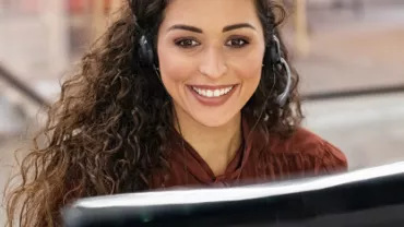 Mulher de cabelos cacheados trabalha em frente ao computador de fones de ouvido e um sorriso no rosto.