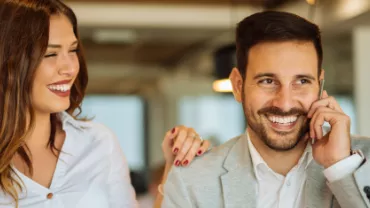 Homem e mulher lado a lado, homem fala ao telefone com sorriso no rosto enquanto mulher celebra