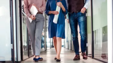 Imagem focada na perna de três pessoas andando pelo corredor de uma empresa enquanto seguram papeis nas mãos. 