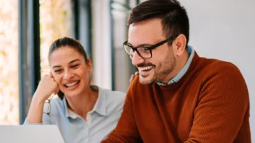 Um homem e uma mulher sentados, sorrindo e olhando para o computador. 