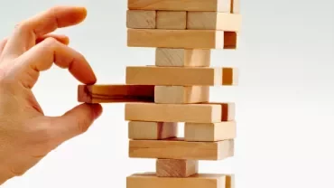 Mão retirando bloco de torre de madeira em jogo de equilíbrio.