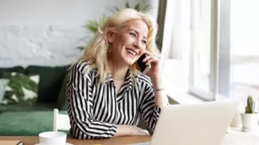 Mulher sorrindo enquanto fala ao telefone, sentada em frente a um laptop.