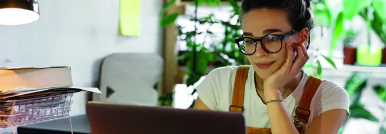 Mulher com óculos trabalha no laptop em uma mesa com plantas ao fundo.