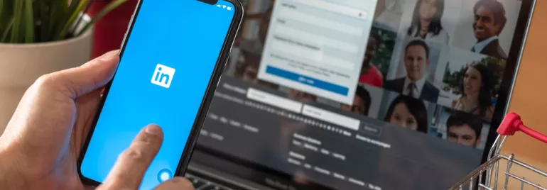 mão segurando celular logado no LinkedIn, notebook logado em LinkedIn ao fundo