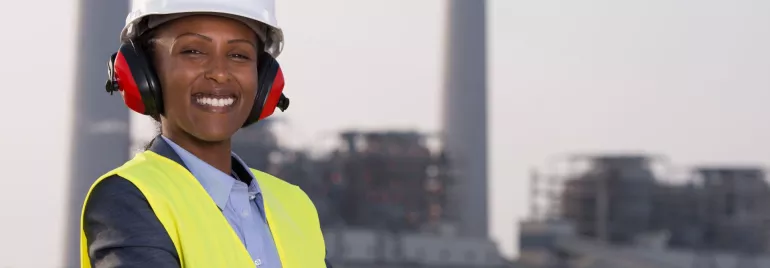 Mulher preta usando equipamentos de proteção na frente de uma indústria