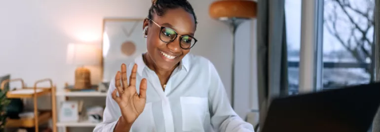 Mulher de pele negra e cabelo preto trançado acena para o computador enquanto está em uma reunião de trabalho.