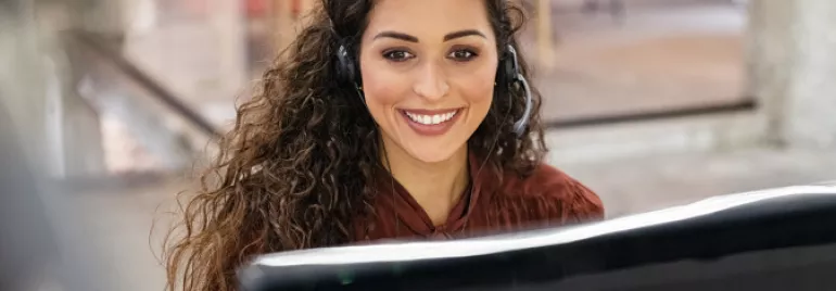 Mulher de cabelos cacheados trabalha em frente ao computador de fones de ouvido e um sorriso no rosto.