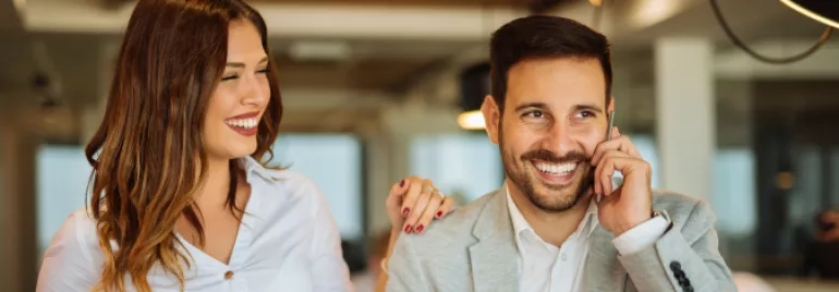 Homem e mulher lado a lado, homem fala ao telefone com sorriso no rosto enquanto mulher celebra