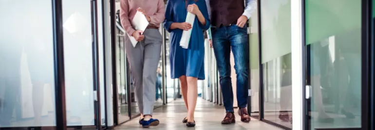 Imagem focada na perna de três pessoas andando pelo corredor de uma empresa enquanto seguram papeis nas mãos. 