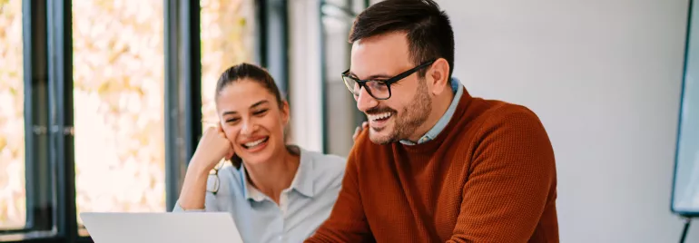 Um homem e uma mulher sentados, sorrindo e olhando para o computador. 