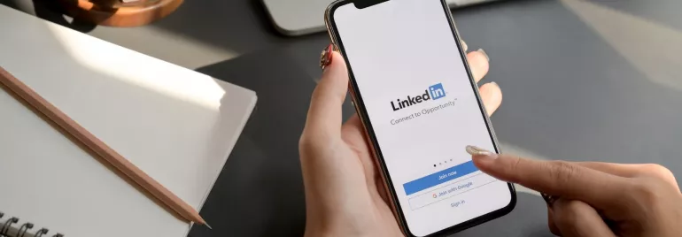 Visto de cima: mesa com cadernos e canetas ao fundo e a mão de uma pessoa segurando um celular com o aplicativo do linkedin