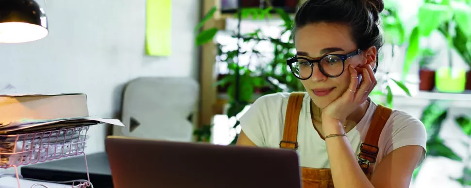 Mulher com óculos trabalha no laptop em uma mesa com plantas ao fundo.