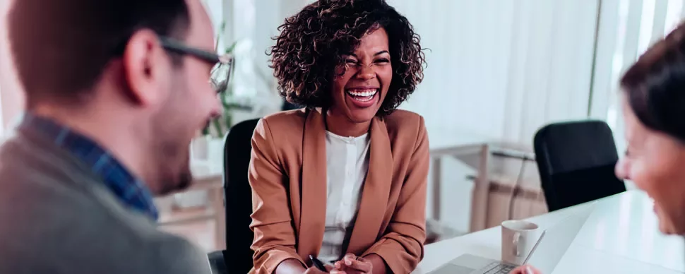 Três pessoas conversando e rindo em uma reunião de trabalho.