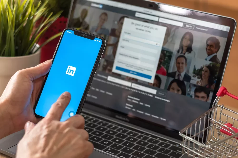 Pessoa usando o app LinkedIn no celular em frente ao laptop.