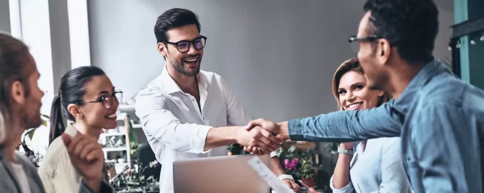 Equipe de trabalho reunida em escritório, duas pessoas apertando as mãos.