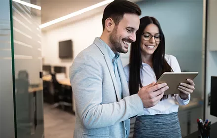 Duas pessoas sorrindo enquanto olham para um tablet em um escritório.