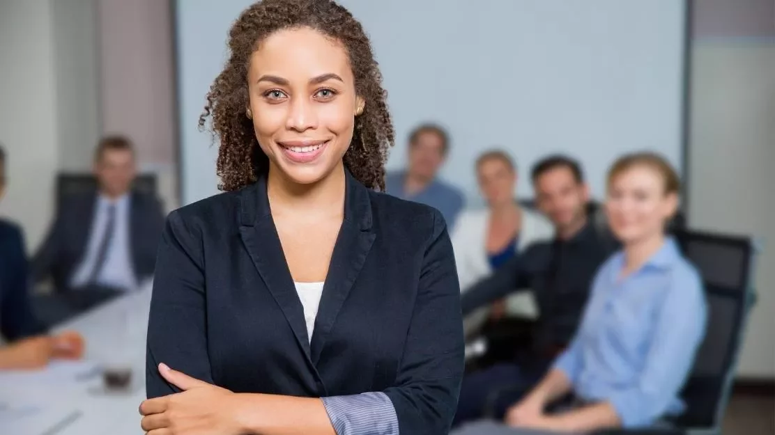 Mulher sorridente em foco, com uma equipe ao fundo em uma sala de reunião.