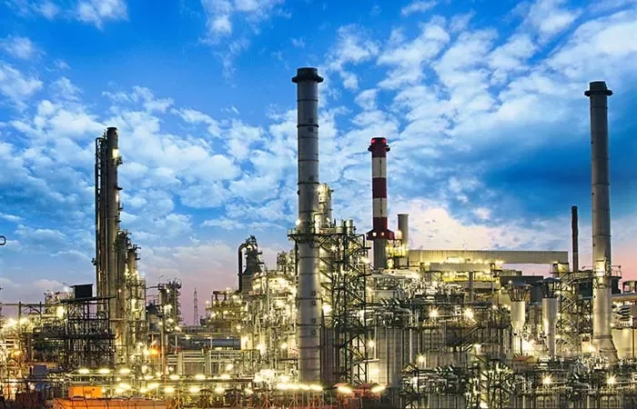 Indústria petroquímica iluminada ao entardecer, com céu azul e nuvens.