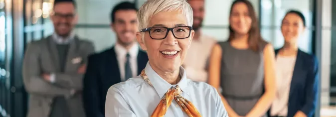 Mulher de cabelos grisalhos sorrindo com equipe ao fundo.
