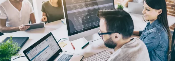 Equipe trabalhando em escritório com computadores e programação na tela.