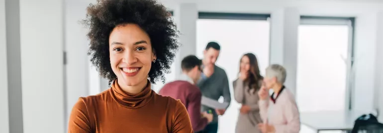 Mulher sorridente em primeiro plano com grupo de pessoas desfocado ao fundo em escritório.