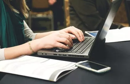 Pessoa digitando em laptop com smartphone e papéis ao lado.