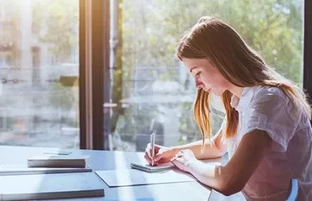 Mulher escrevendo em um caderno, iluminada pela luz do sol através da janela.