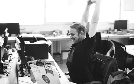 Homem sorridente se espreguiça em frente ao computador em um escritório.