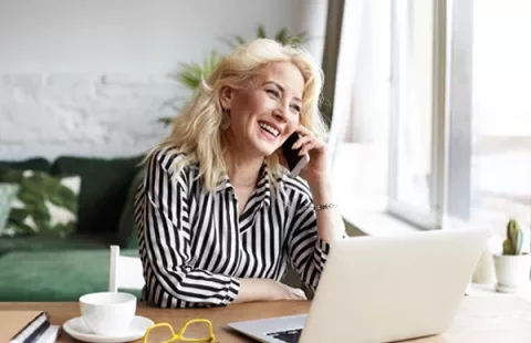 Mulher sorrindo enquanto fala ao telefone, sentada em frente a um laptop.