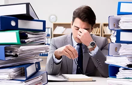 Homem cansado no escritório cercado por pilhas de pastas azuis.